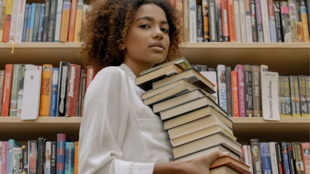 Young woman in a library holds a large stack of books that lean against her body.
