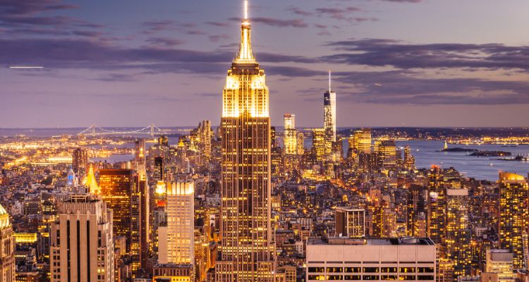 The image depicts the skyline of New York City with the Empire State Building towering over the other buildings. The city is bathed in gold light from all the buildings lit up at night.