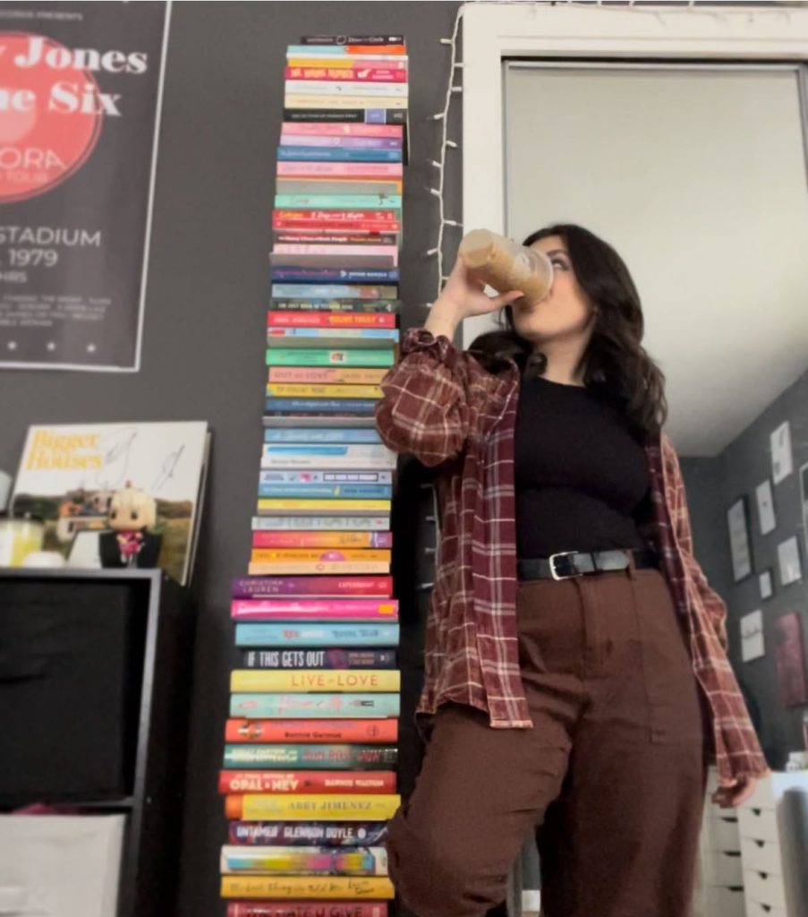 Bookstagrammer Lindsey with her hip popped, drinking an iced latte, with a towering stack of books going over her head in her room decorated with vinyls and posters.