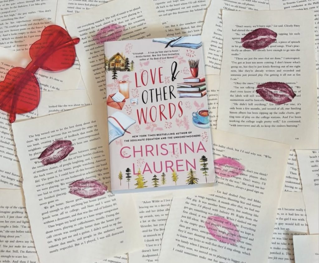Romance book cover Love & Other Words by Christina Lauren on top of loose pages with pink and purple kiss stains on them. In the corner is a pair of red heart sunglasses.