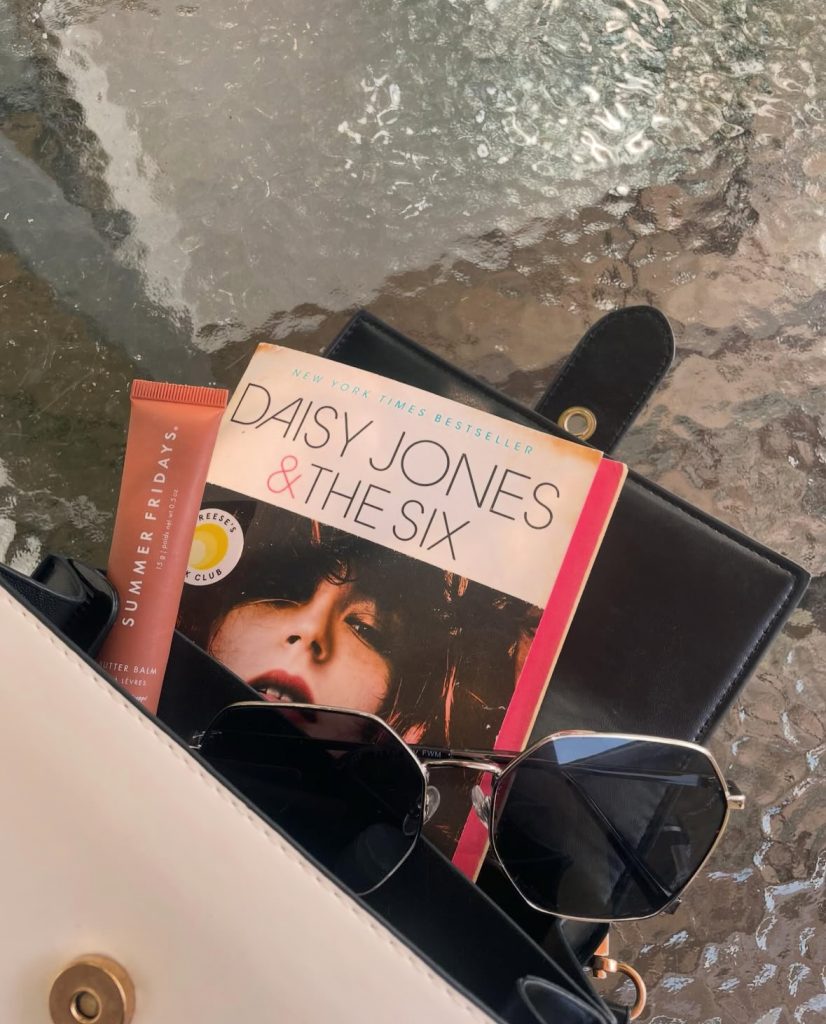 On top of a glass table is Bookstagrammer Lindsey's purse with a peach lipgloss, black sunglasses, and Daisy Jones & The Six by Taylor Jenkins Reid spilling out.