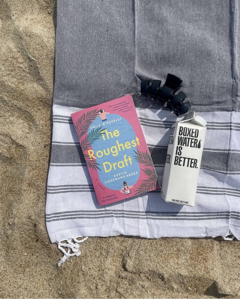 On a sandy beach on top of a towel sits pink and blue fiction book cover of The Roughest Draft by Austin Siegemund-Broka, a black claw clip, and a boxed water.