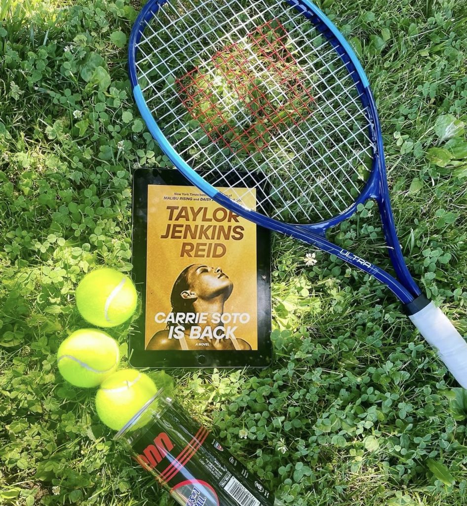 A kindle laying outside in the grass with the golden book cover Carrie Soto Is Back by Taylor Jenkins Reid. Next to it is a blue tennis racket and three yellow tennis balls falling out of their packaging.