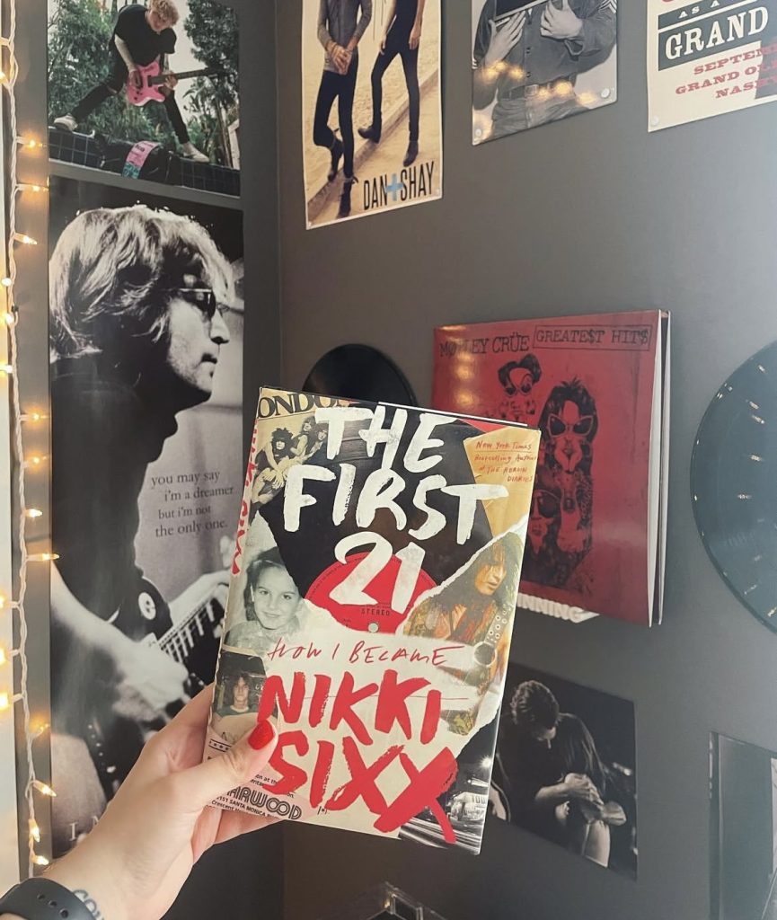 Bookstagrammer Lindsey holding up the nonfiction book The First 21: How I Became Nikki Sixx in front of a black wall lined with rock posters (including John Lennon), records, and twinkly lights.