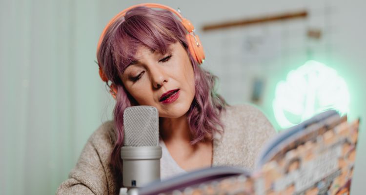 The image is of a woman with pinkish hair and bangs with orange headphones reading out loud with an open book and microphone in front of her.