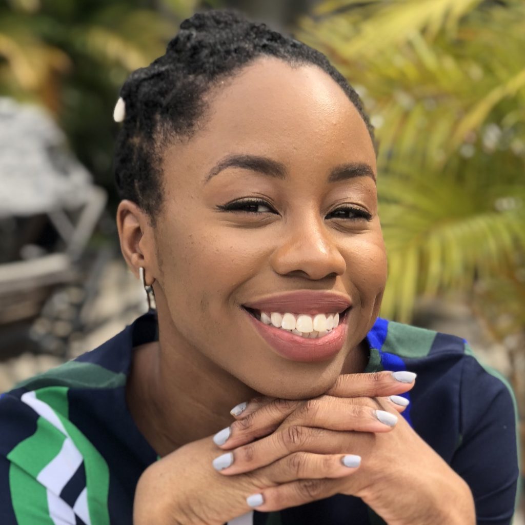 A'rese Emokpae, a Black woman with braided hair tied back, smiles at the viewer while resting her chin on her hands. Large leaves can be seen in the background.