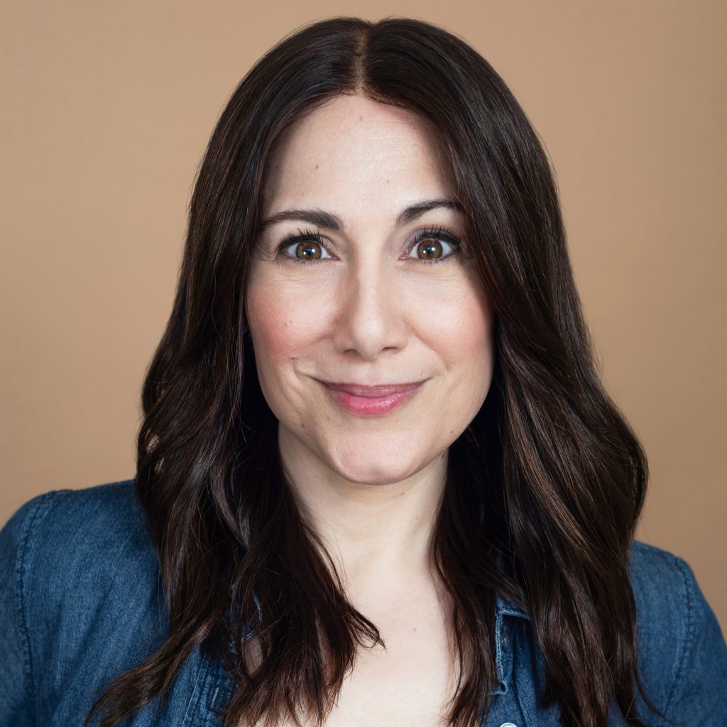 Amy Hutchins, a light-skinned woman with long dark brown hair and a denim shirt, smiles at the viewer against a light brown background.