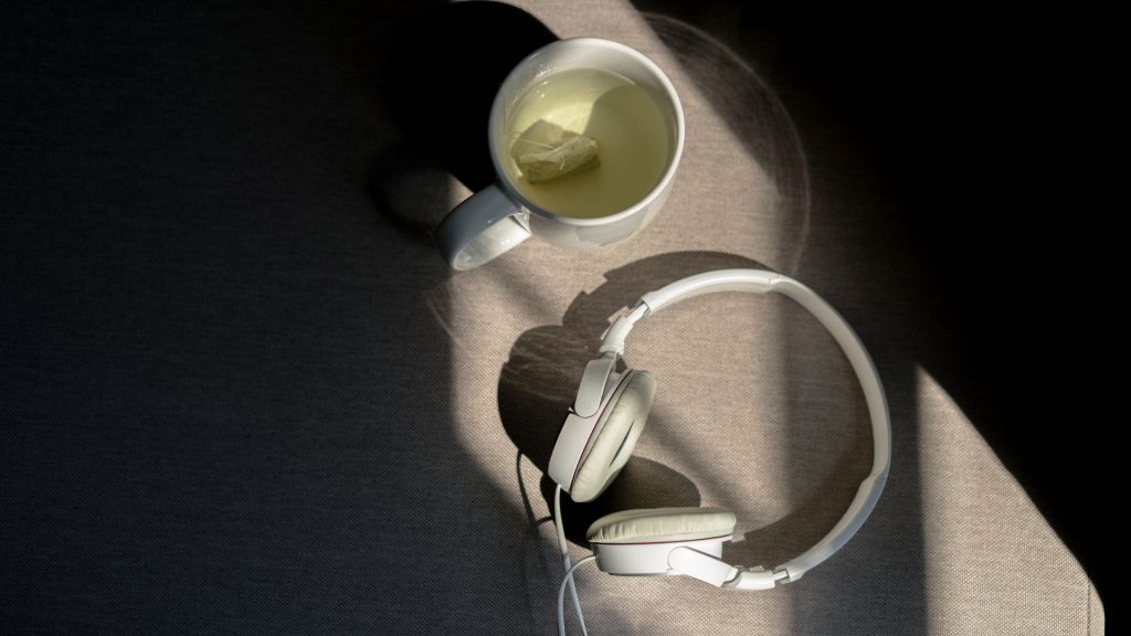 A mug of tea and a pair of white headphones sit on a table, illuminated by a narrow strip of light cutting through the darkness.