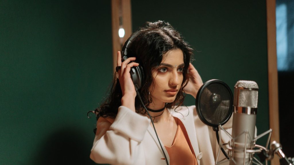 A young woman with light brown skin, dark wavy hair, and a bindi on her forehead puts on a pair of headphones as she stands in front of a mic in a green-walled recording booth.