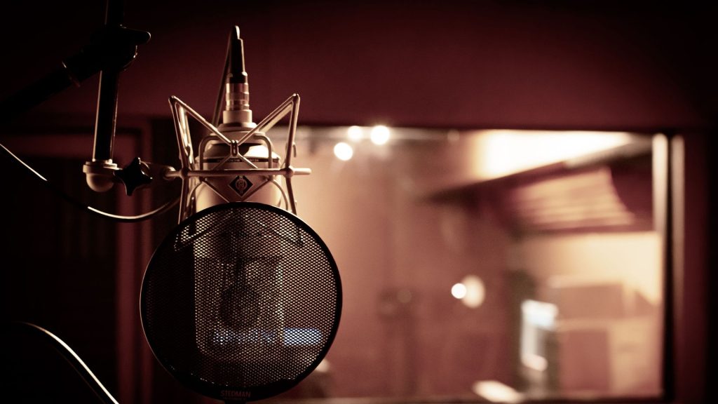 A microphone and pop filter inside an empty, dimly lit recording booth with the control room visible through the window.