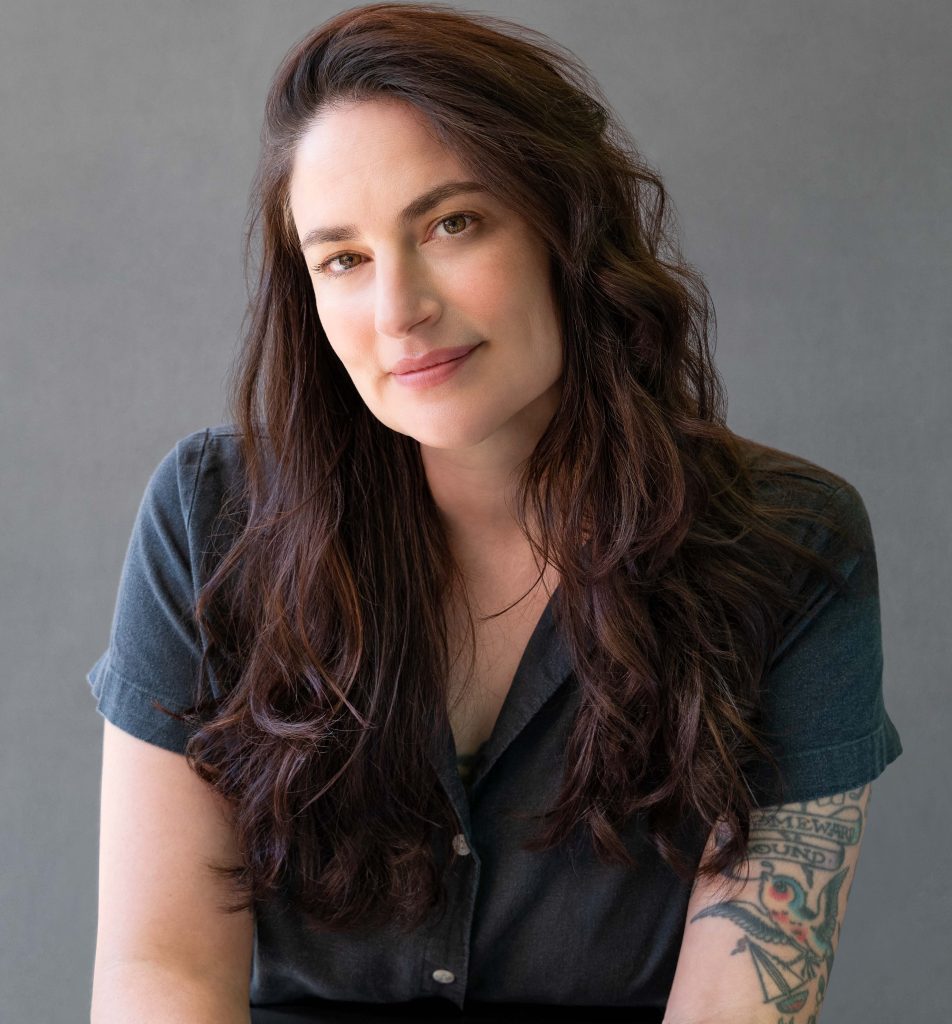 Shoshana von Blanckensee, a light-skinned woman with long dark hair and a tattoo sleeve on her left arm, looks at the viewer. She's wearing a blue short-sleeve shirt, and the wall behind her is gray.