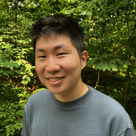 Andy Jiaming Tang, a Chinese man with short black hair and a gray shirt, smiles at the viewer. He's standing in front of an array of trees.