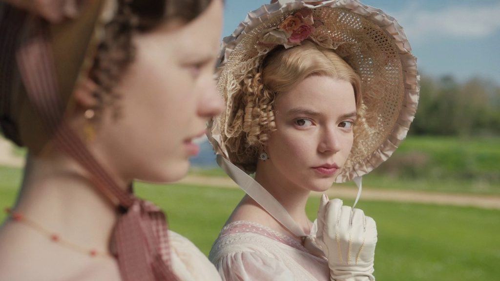 Image from Emma (2020) of Emma looking at her Harriet, who is out of focus. She is wearing a straw bonnet with pink accents.