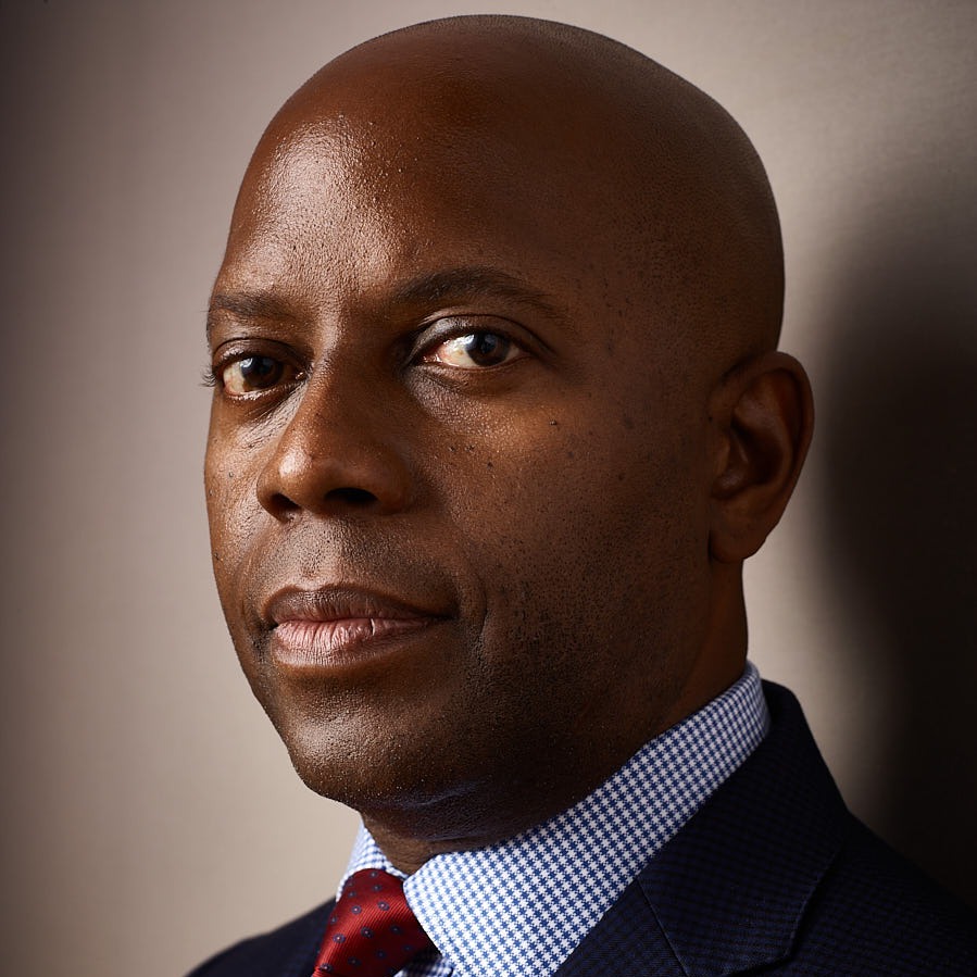 Dr. Brian H. Williams, a bald Black man in a suit, looks at the viewer from a three-quarter angle facing left against a beige background.