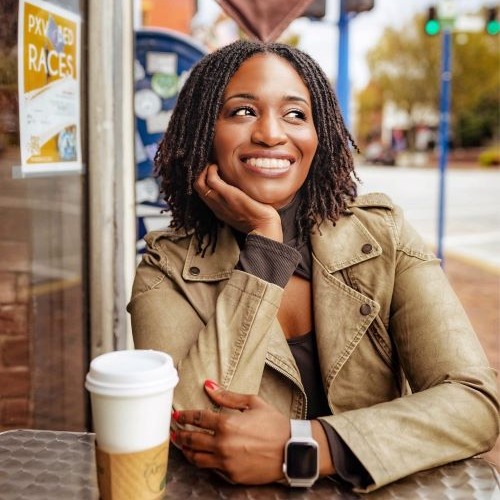 Khristi Lauren Adams, a Black woman in a khaki jacket, smiles as she looks off to the right and props her chin on her hand. She is sitting at a table outside a cafe with a coffee cup beside her elbow.
