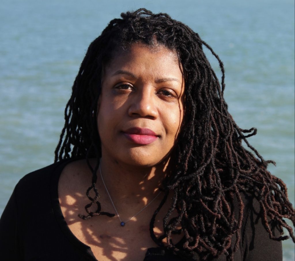 Mikki Kendall, a Black woman with locs and a black shirt, looks at the viewer with the ocean in the background.