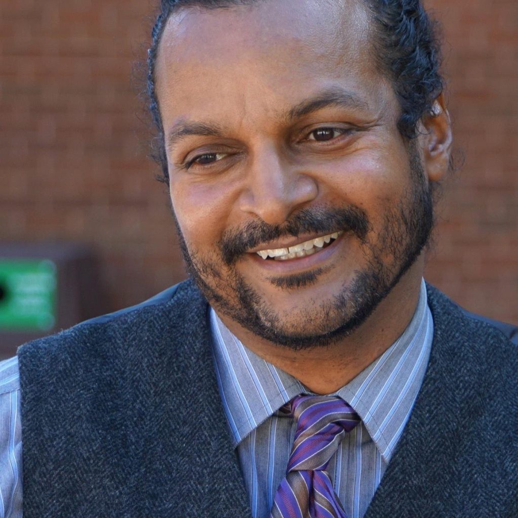 Tony Medina, a Black man with facial hair in a dress shirt, tie, and vest, looks off to the bottom left as he smiles. Out of focus behind him is a brown wall.