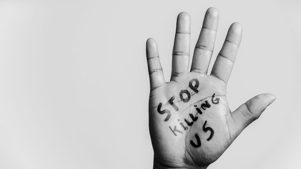 A hand with the phrase "Stop killing us" written in marker on the palm is shown in black and white.