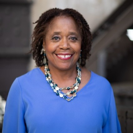 Dr. Marietta Collins, a Black woman in a blue shirt and necklace, smiles at the viewer with a building out of focus in the background.