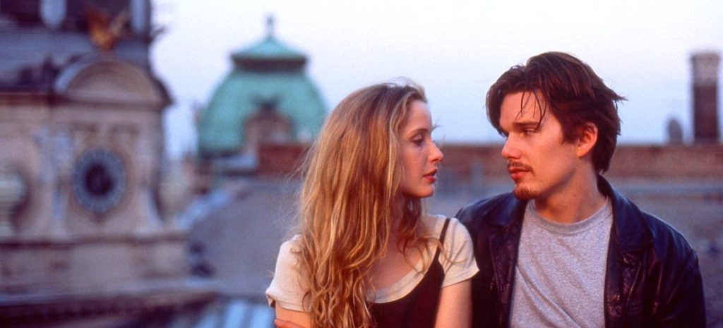 A still from the 1995 movie Before Sunrise. A young blonde woman in a white t-shirt and black tank top dress stares into the eyes of a young brunette man in a gray t-shirt and black leather jacket. Behind them are beautiful classical buildings with white stone and green domed roofs. The two are lit by the pinkish-orange haze of a sunset.