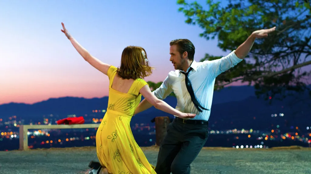 A still from the 2016 movie La La Land. A woman in a yellow dress and a man in a white button down shirt, black tie, and black pants are poised mid-tap dance with their arms splayed out to the side. Behind them is the Los Angeles skyline shaded by a pale pink, purple, and blue sunset.
