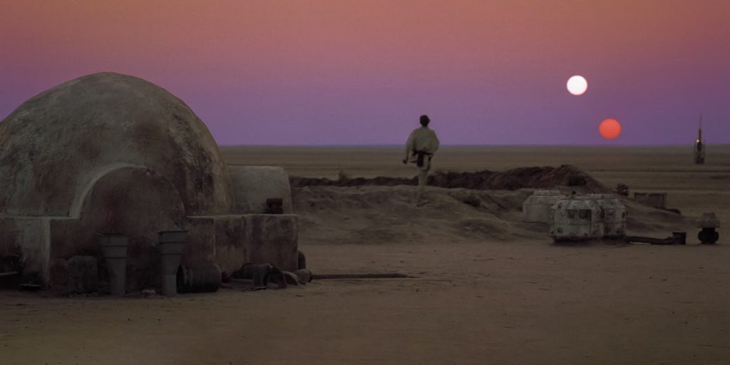A still from the 1977 movie Star Wars: Episode IV — A New Hope. In a desert landscape with a domed stone house, a young man in a pale tunic and pants walks off toward a purple sunset. Two suns hang in the sky: one white, the other a reddish orange.