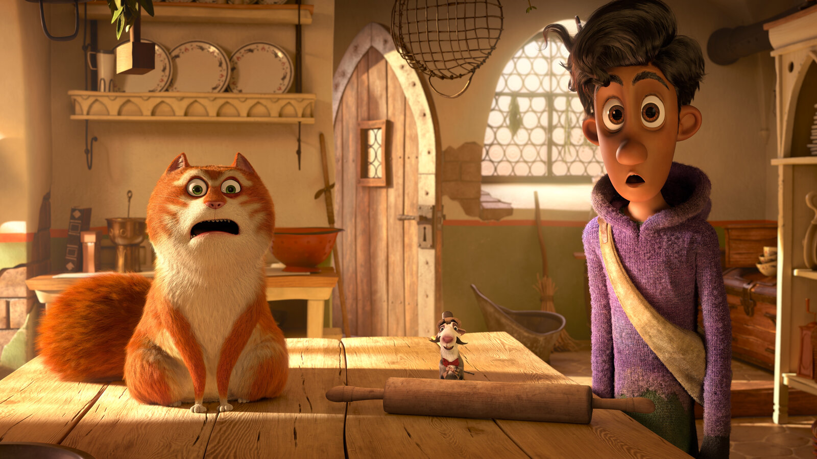 Movie still from Narrativia's 'The Amazing Maurice'. Image depicting a large orange and white cat with a big bushy tail sitting on a wooden table, staring confusedly at a moustachioed rat wearing a brown top-hat. The room behind them appears to be a kitchen, with colander and other cooking utensils on another table behind them. A wooden door stands half-way open to the right of the frame. A young man in a purple hooded jumper with brown skin, eyes, and dark hair stands with his mouth open to the left of the rat. He has a beige cross-body bag across his chest.