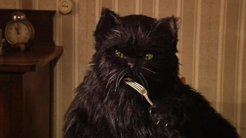 Movie still from Mars Media Entertainments adaptation of 'The Master and Margarita'. Image depicting a large black cat with sharp claws, white whiskers and yellow-green eyes eating food from a fork. He sits on a chair in front of brown wallpapered walls with a wooded side-table to his left behind him, on which sits a bronze alarm clock reading 11 o'clock.