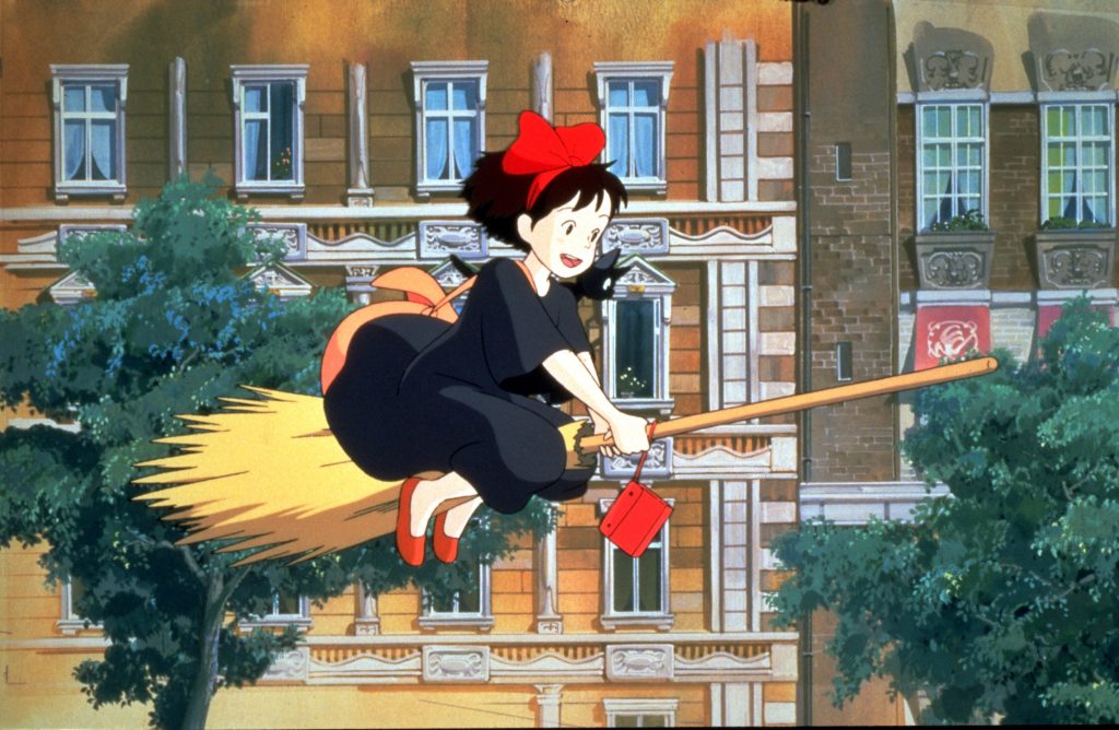 Movie still of Kiki's Delivery Service, a young girl in a dark black dress is flying on a broom, passing buildings nearby. She is wearing red shoes, a red bow, and a black cat is sitting on her shoulder, peering down below.