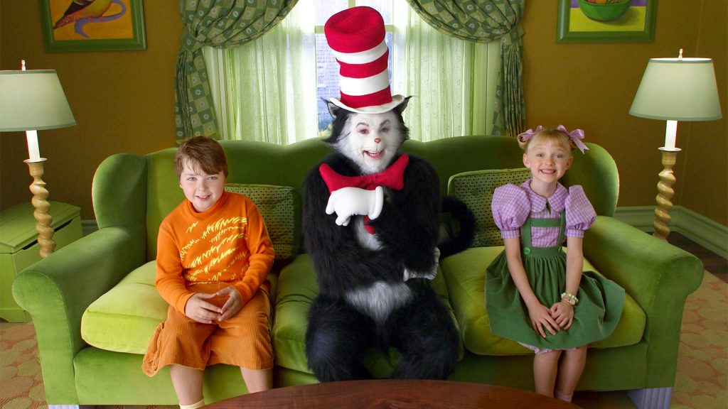 Movie still from Universal Pictures' The Cat in the Hat. Image depicting a large anamorphous white and black cat wearing a big red and white top hate and red bow tie. He sits on a push green sofa in the middle of two children, a boy and a girl. The boy to his left is wearing an orange graphic jumper and orange shorts. He has brown hair swept over to the right of his face. The girl on the right has blonde hair in two pigtails with pink bow ribbons. She is wearing a white and lilac blouse and dark green pinafore dress. The sofa is placed in front of a large sitting room window with patterned green curtains. A white lamp stands at either end of the sofa.