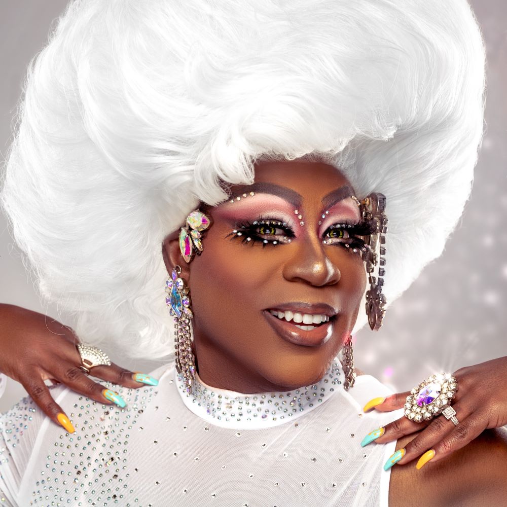 Felicia "Fefe" Minor, a smiling brown-skinned drag queen wearing vibrant green contact lenses, a towering white bouffant wig and white rhinestone blouse, yellow and blue nails, and various pieces of costume jewelry and gemstones.
