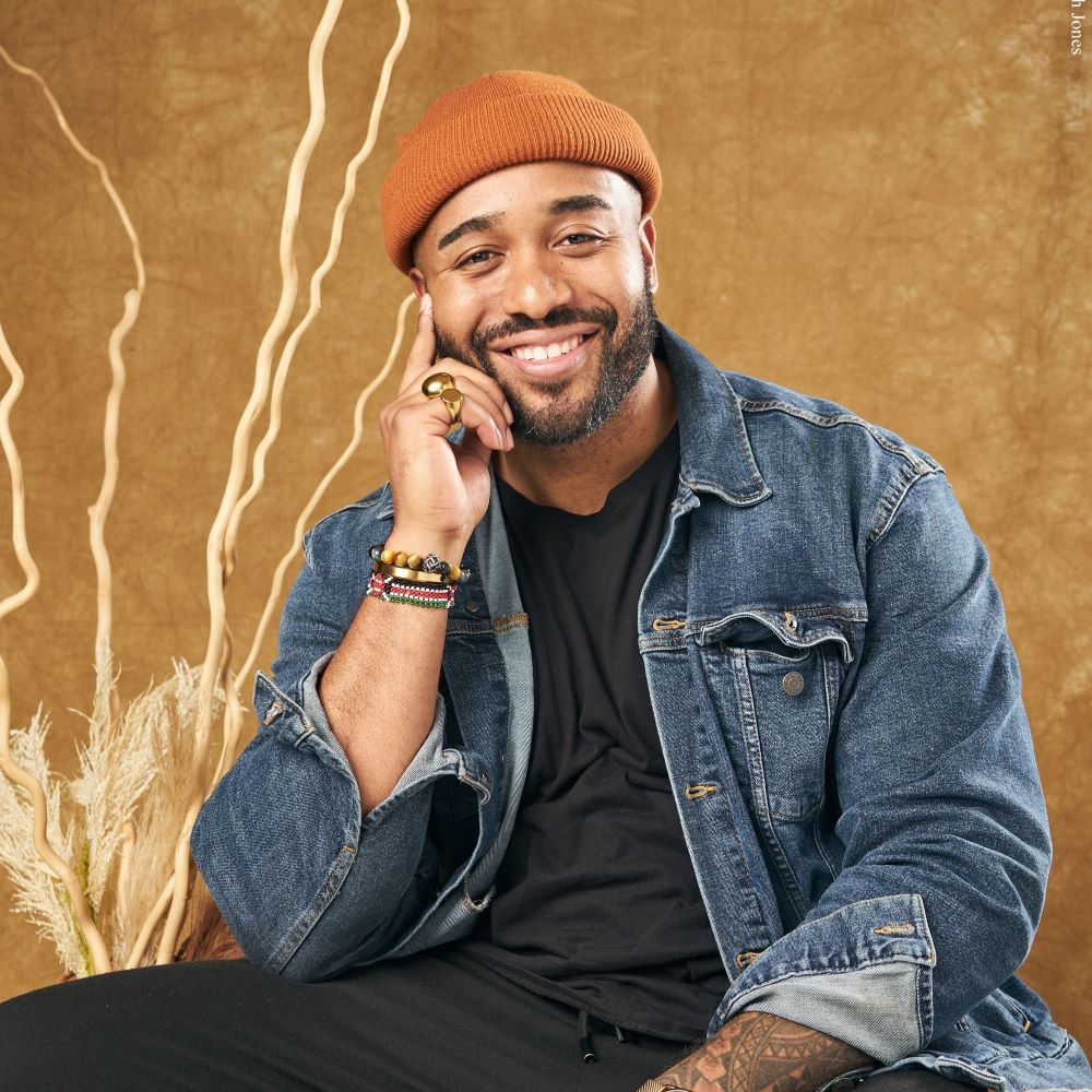 Frederick Joseph, a smiling brown-skinned, brown-eyed man with black mustache and beard, wearing an orange beanie, black t-shirt and pants, and a denim jacket, as well as gold rings and a collection of bracelets on one hand, against a tan background.