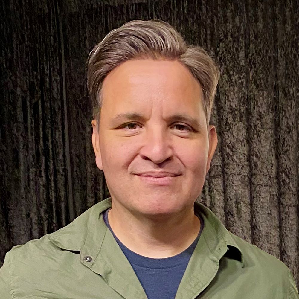 John Parra, a tan-skinned man with short, straight salt-and-pepper hair and brown eyes, wearing a green windbreaker over a blue t-shirt, posing against a patterned background.