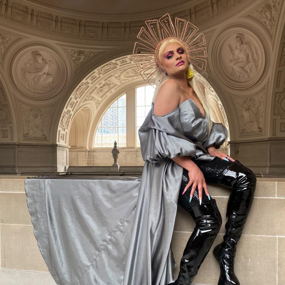 Per Sia, a light-skinned drag queen with a long, straight blonde wig, wearing a draping silver sleeveless dress over a thigh-high pair of black books and black sequined leggings, a golden wire headdress, and large yellow earrings.