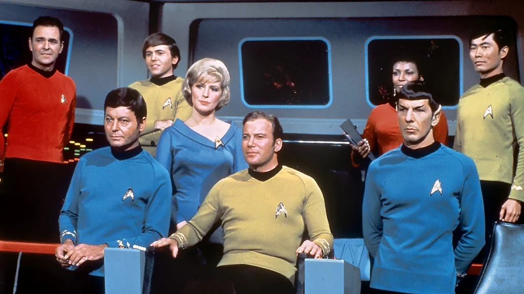 Still image from the television show Star Trek: The Original Series, featuring a group of adults diverse in skin tone and hair color/texture, wearing coordinating uniforms on a spaceship's command deck.