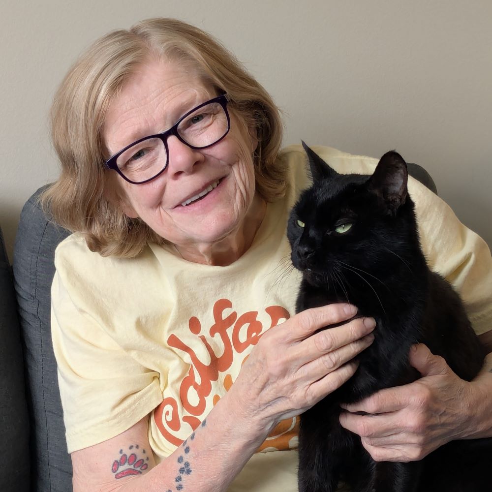 Tamora Pierce, a smiling elderly light-skinned woman with shoulder-length, wavy blonde hair and eyeglasses, wearing a yellow t-shirt and with visible cat paw tattoos on her forearm, holding a shorthair black cat in her lap while seated on a gray chair.