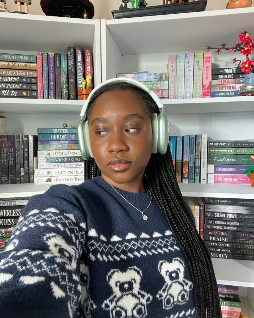 Darmen of @darmensbookshelf, a young Black woman with box braids, white headphones, and a blue and white sweater, poses in front of a white bookshelf looking off to the viewer's left.