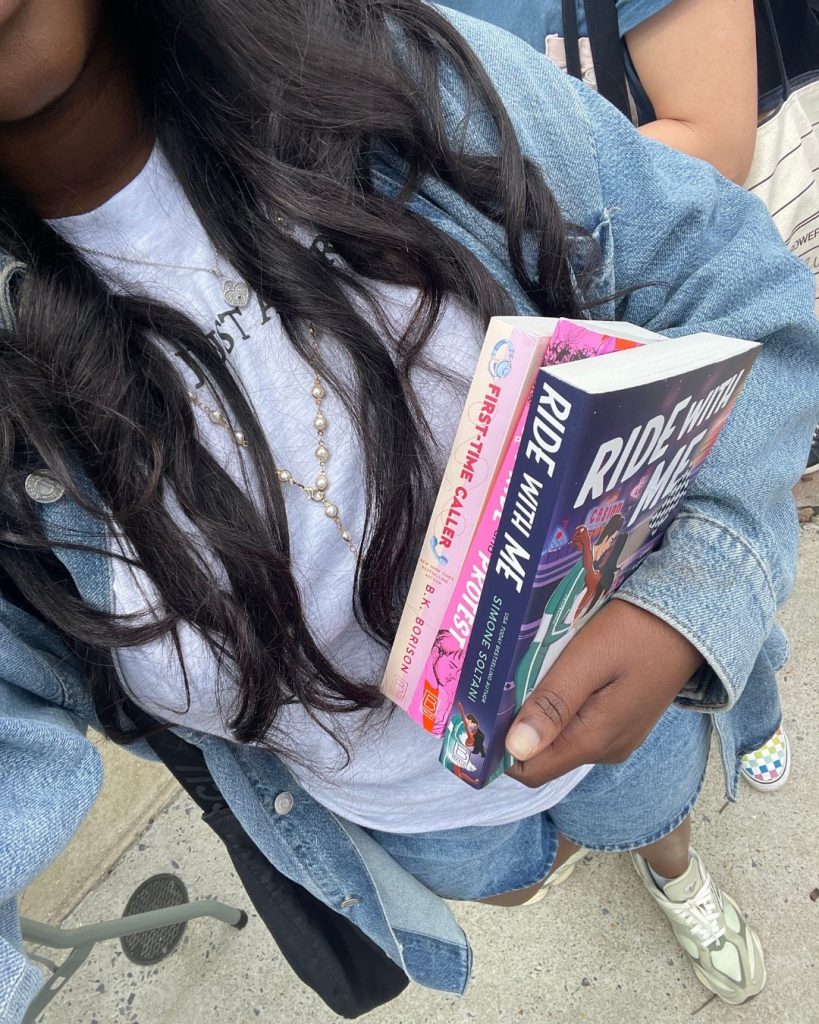 Darmen (@darmensbookshelf) holds copies of First-Time Caller by B.K. Borison, Pride and Protest by Nikki Payne, and Ride with Me by Simone Soltani. She's standing on pavement outside and people can be seen behind her in line.