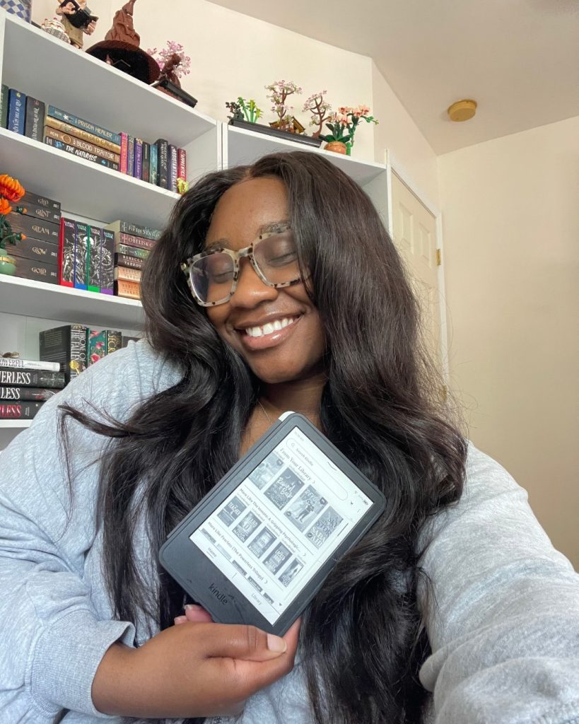 Darmen of @darmensbookshelf holds a Kindle in her right hand while smiling with her eyes closed. She wears a gray sweatshirt and oversized glasses, and her hair is long and straight. A white bookshelf can be seen behind her.