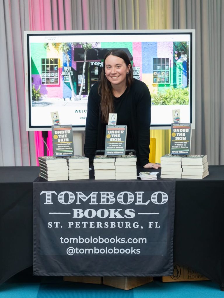 A woman wearing a black long sleeved top stands behind a counter and stacks of books. A black banner is placed in front of the counter and a picture of the colorful bookstore is in the background.