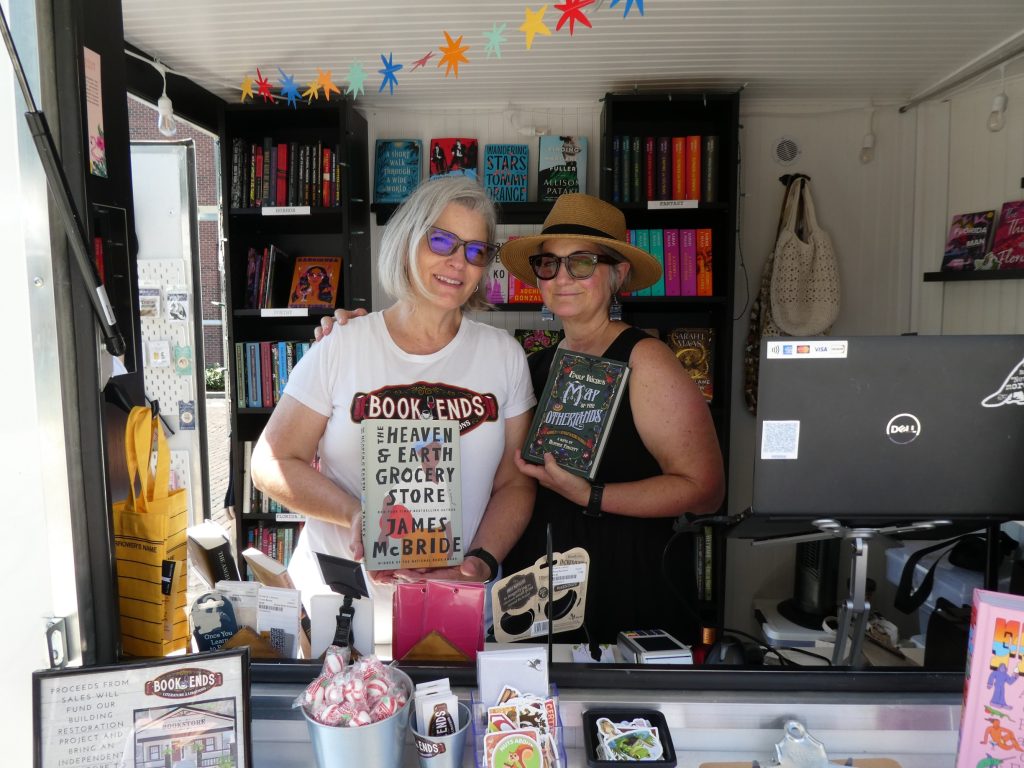 Two women wearing sunglasses are standing in a trailer filled with books and other items. They are both holding books.
