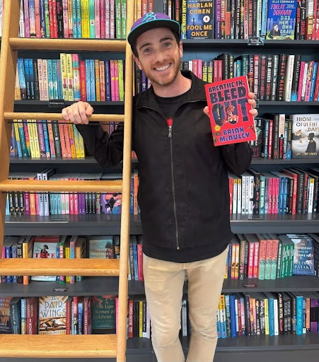 Author Brian McAuley, White man smiling holding up book whilst beside bookshelves with rolling ladder.