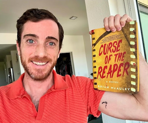 White man, Brian McAuley, author, smiling wearing a bright coral top. Holding up yellow book 'Curse of the Reaper', small scythe tattoo on his arm.
