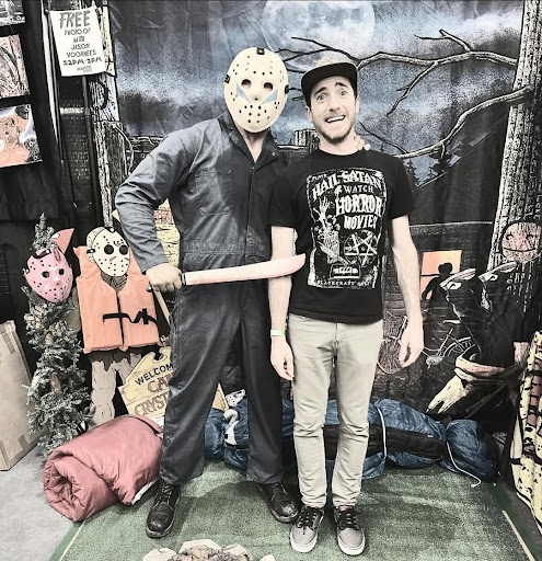 White man posing in shock whilst held by horror character Jason Vorhees. Horror movie memorabilia in photo's frame.