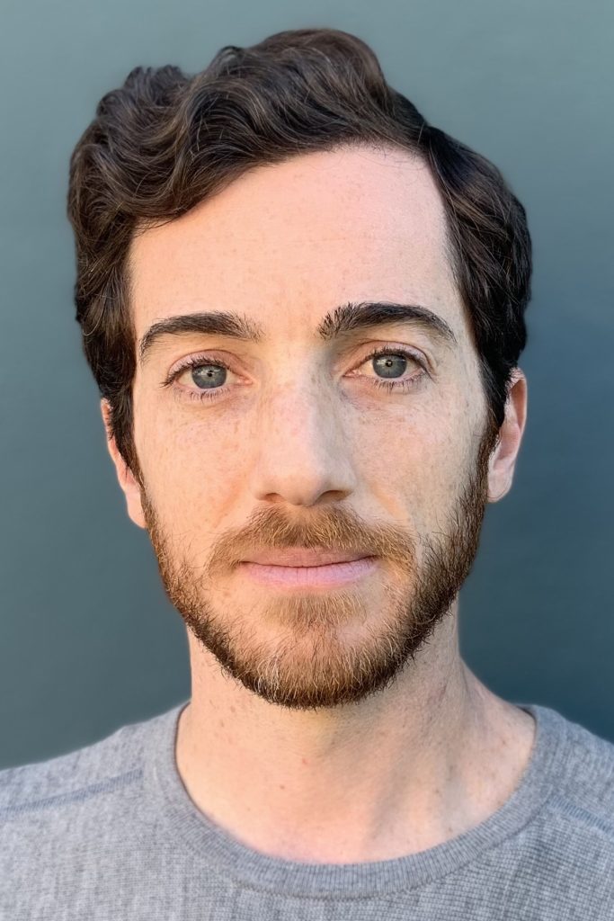 Author Brian McAuley, a white man with a beard, blue eyes and brown hair looking at the camera, standing in front of blue background
