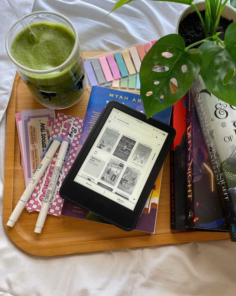A flatlay on a white sheet with tab-sized Post-It notes, two pens, stationery, a Kindle, stacked books, a glass of green juice, and a potted plant all arranged on a wooden tray.