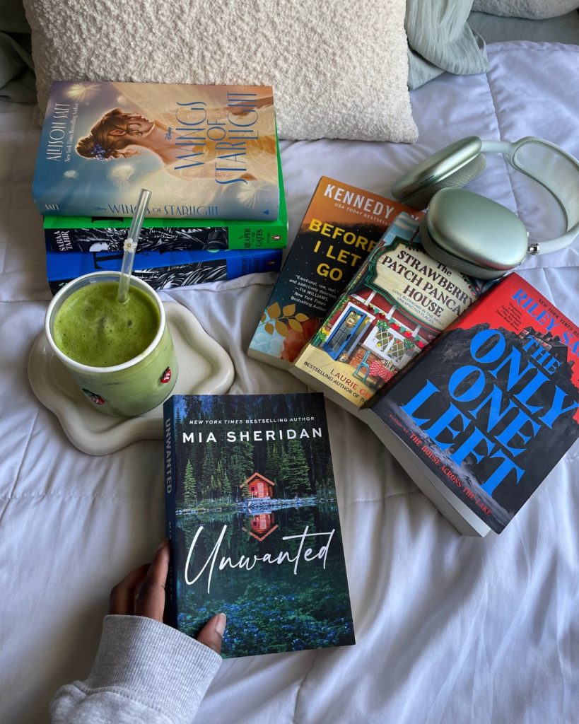 A flatlay with a pillow and several books arranged on a white sheet, along with a pair of green headphones and a glass of green juice with a straw on a small tray. Darmen's hand can be seen in the bottom of the frame, touching a copy of Unwanted by Mia Sheridan.