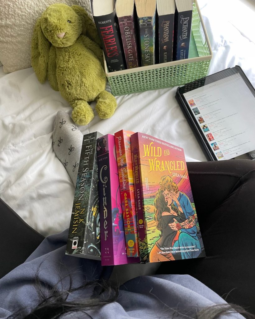 Top-down view of Darmen (@darmensbookshelf) with four books stacked in her lap as she sits atop a white sheet. In front of her is a stuffed green bunny, a basket holding five books, and an open Kindle.