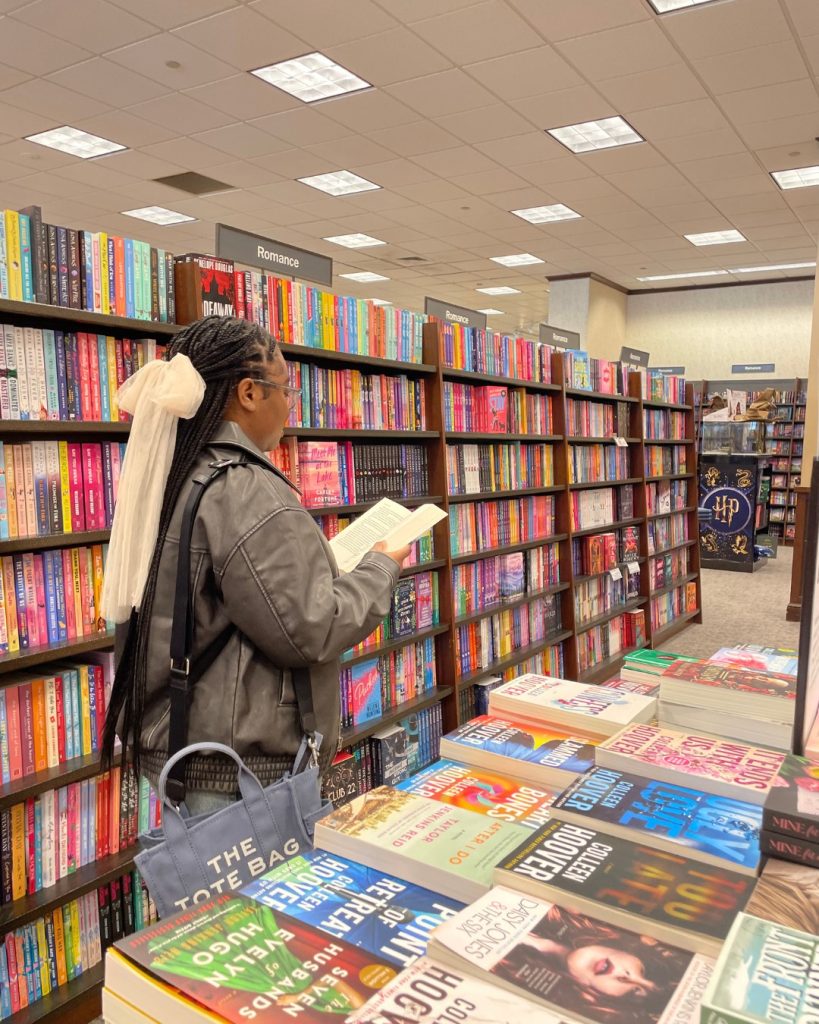 Darmen of @darmensbookshelf stands between a bookshelf and a book display at Barnes and Noble, reading a book. She is wearing a gray jacket and has a sheer pink bow in her hair.