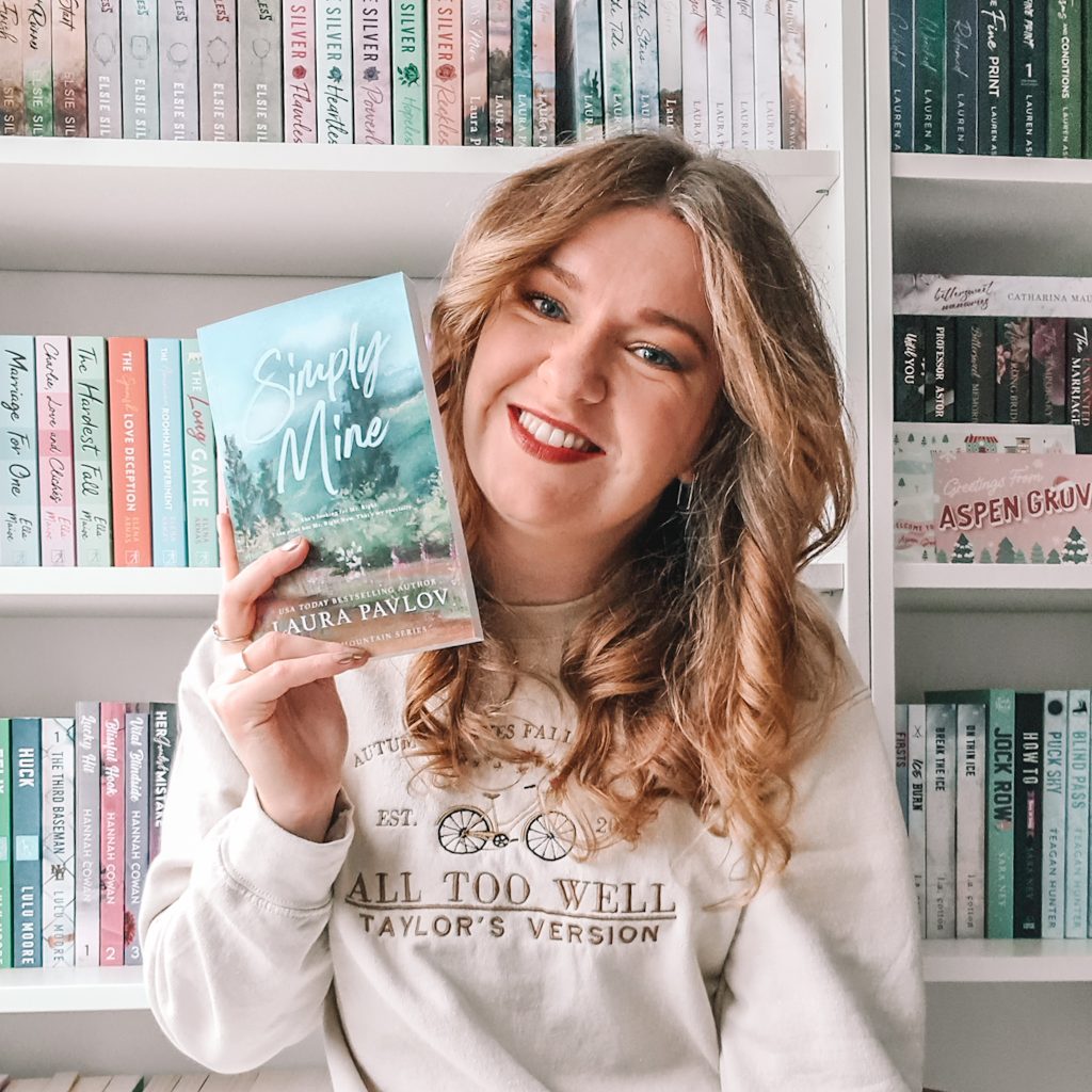 Charlotte of @charlottes.loveforbooks, a light-skinned woman with long blonde hair, smiles at the viewer as she holds up a copy of Simply Mine by Laura Pavlov in front of a white bookshelf packed with books.